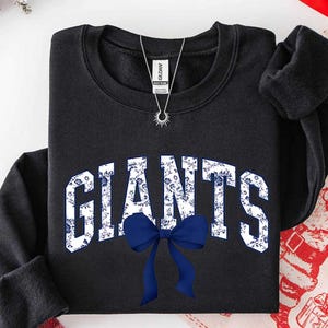 May include: A black crewneck sweatshirt featuring the word GIANTS in large, white, floral-patterned block letters across the chest. A solid navy blue bow is positioned directly below the text. A silver sunburst pendant necklace rests at the neckline.