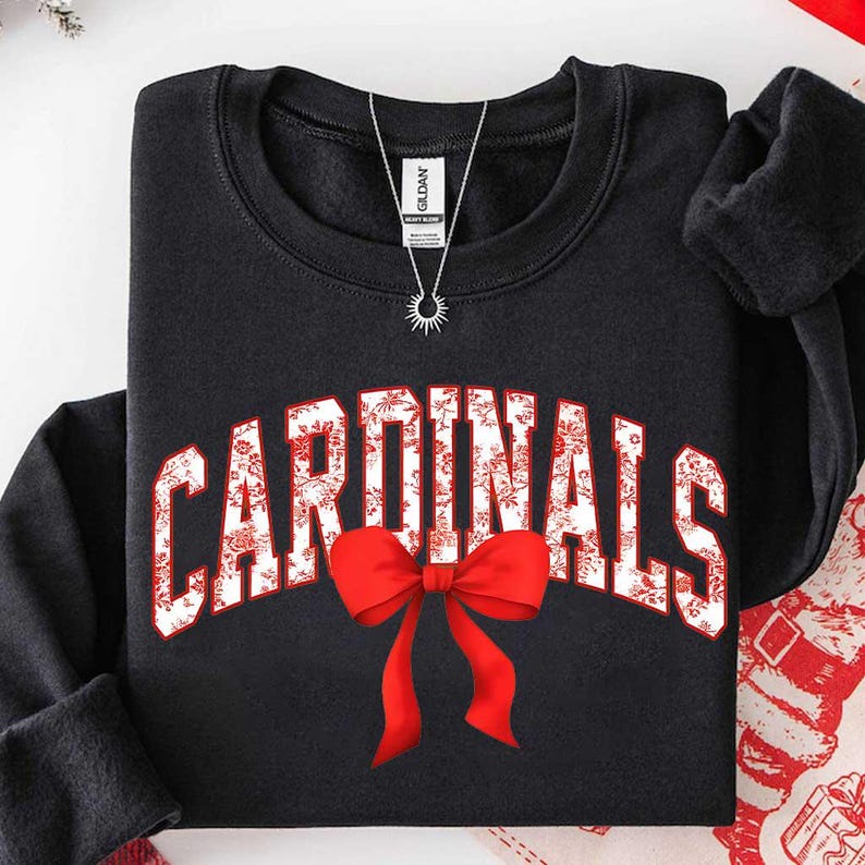 May include: A black long-sleeve sweatshirt featuring the word CARDINALS in white block letters with a red floral pattern. A large, bright red bow is positioned directly below the text. A silver chain necklace rests on the neckline of the shirt.