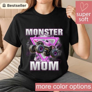 May include: Black t-shirt featuring a pink monster truck design with the words "MONSTER MOM" in white and pink. The truck is set against a purple and black background, with the words "more color options" at the bottom.