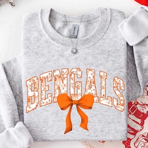 Op de afbeelding: Een lichtgrijs sweatshirt met ronde hals en het woord BENGALS in een wit lettertype met een oranje bloemenpatroon. Onder de tekst is een oranje strik geplaatst. Het kledingstuk heeft een geribbelde kraag, boorden en een zichtbaar merklabel.