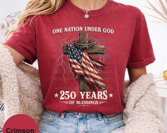 May include: Crimson-colored t-shirt with the text "ONE NATION UNDER GOD" above a wooden cross draped with an American flag. The shirt also features the text "250 YEARS OF BLESSINGS".