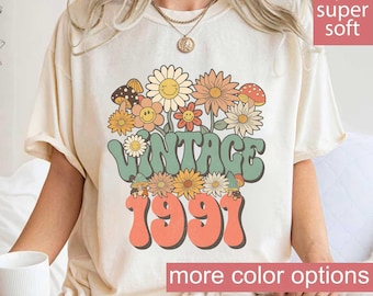 May include: Ivory-colored t-shirt with a retro floral design and the text "VINTAGE 1991." The graphic features smiling flowers and mushrooms in shades of orange, yellow, and green. The shirt has a relaxed fit and short sleeves.