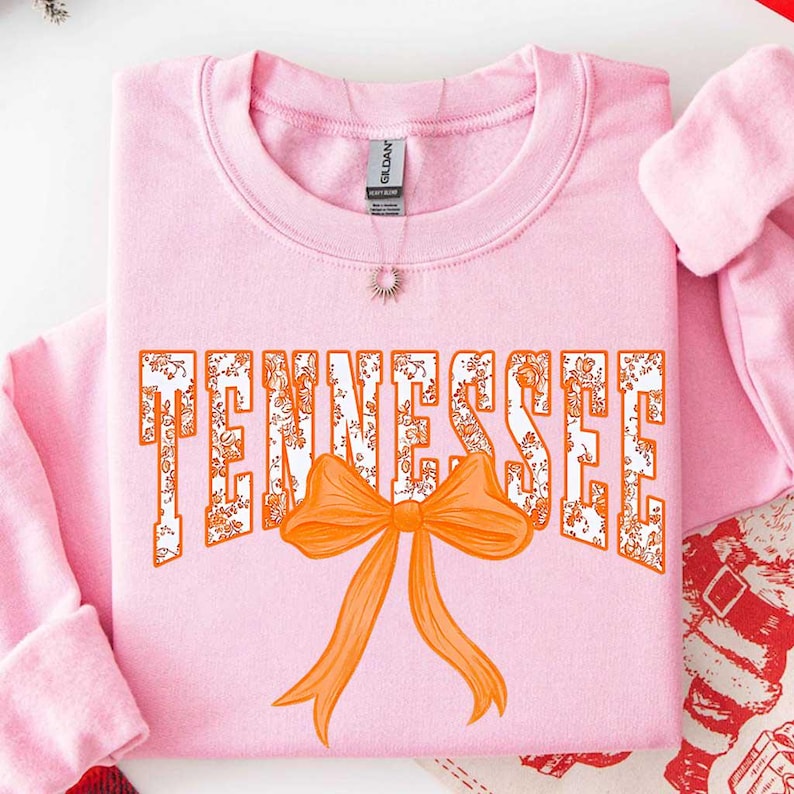 K&ouml;nnte beinhalten: Ein hellrosa Sweatshirt mit Rundhalsausschnitt, das das Wort TENNESSEE in gro&szlig;en, wei&szlig;en Blockbuchstaben mit einem orangefarbenen Blumenmuster zeigt. Unter dem Text befindet sich eine gro&szlig;e, einfarbig orangefarbene Grafik einer Schleife.