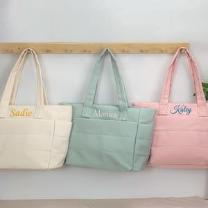 May include: Three quilted tote bags in cream, sage green, and light pink. Each bag features two shoulder straps, side pockets, and a horizontal quilted pattern. The cream bag is embroidered with the name Sadie, the green with Monica, and the pink with Kaley.