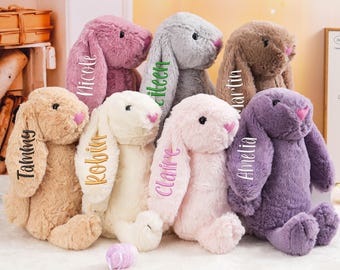 May include: A collection of plush bunny rabbits in various pastel colors, including pink, purple, beige, and gray. Each bunny has a name embroidered on its ear. The bunnies have pink noses and are designed for children.