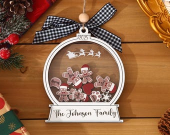 May include: A white wooden Christmas ornament with a clear center, featuring gingerbread people and a Santa sleigh design. The ornament has a black and white checkered bow and reads "2025" and "The Johnson Family."