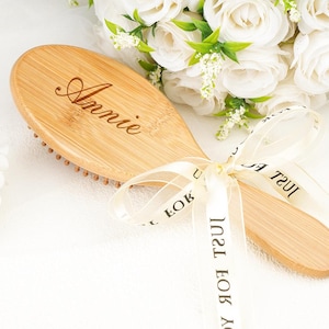 May include: A wooden hairbrush with the name "Annie" engraved on the handle. The brush is tied with a cream-colored ribbon that has text printed on it. White roses and greenery are in the background.