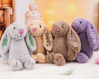 May include: Four plush bunny rabbits in gray, tan, brown, and purple. Each bunny has a pink nose and personalized text on the ear. The bunnies are sitting on a white surface, with Easter eggs in the background.