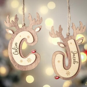 May include: Two wooden reindeer-shaped Christmas ornaments hanging from twine. One ornament features the letter C with the name Colin, and the other features the letter J with the name Jacob. Both have small red noses and snowflake engravings.