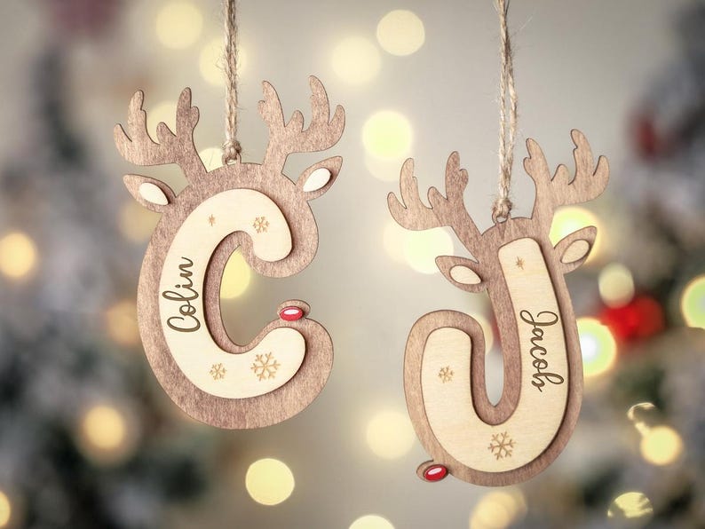 May include: Two wooden reindeer-shaped Christmas ornaments hanging from twine. One ornament features the letter C with the name Colin, and the other features the letter J with the name Jacob. Both have small red noses and snowflake engravings.