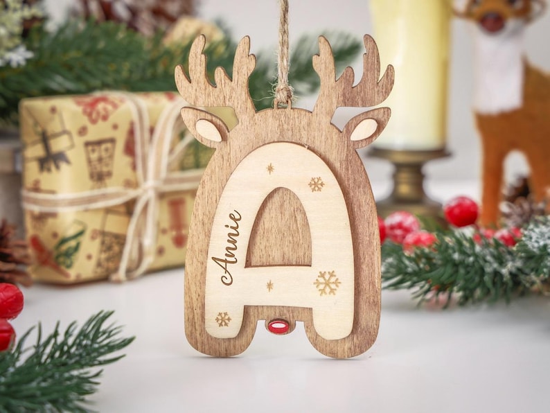 May include: A wooden Christmas ornament shaped like a reindeer head with a layered light-colored letter A in the center. The letter is engraved with the name Annie and small snowflake designs. A small red accent is visible at the base of the letter.