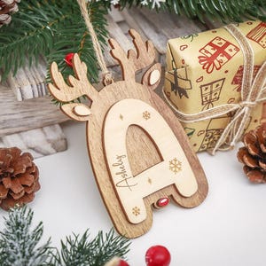 May include: A wooden Christmas ornament shaped like a reindeer head with a layered letter A in the center. The letter A is engraved with the name Ashely and small snowflake designs. A jute twine loop is attached to the top for hanging purposes.