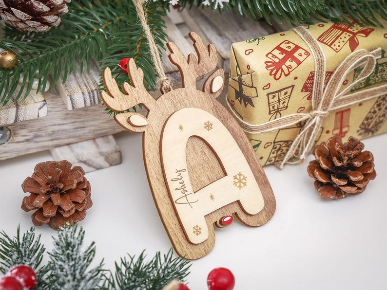 May include: A wooden Christmas ornament shaped like a reindeer head with a layered letter A in the center. The letter A is engraved with the name Ashely and small snowflake designs. A jute twine loop is attached to the top for hanging purposes.