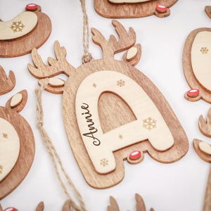May include: A wooden letter ornament shaped like a reindeer with antlers and a red nose. The center features a lighter wood inlay with the name Annie engraved in black script, surrounded by small engraved snowflake patterns and a jute twine loop.
