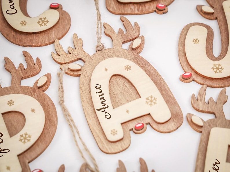 May include: A wooden letter ornament shaped like a reindeer with antlers and a red nose. The center features a lighter wood inlay with the name Annie engraved in black script, surrounded by small engraved snowflake patterns and a jute twine loop.