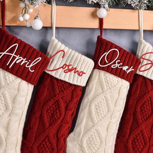 May include: Four personalized Christmas stockings hang from a wooden beam. Each stocking is red and cream-colored with a cable knit design. The names "April", "Cosmo", "Oscar", and "Simon" are embroidered in white script on the cuffs. Decorative greenery and ornaments are visible.
