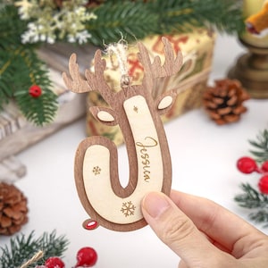 May include: A wooden Christmas ornament shaped like the letter J with reindeer antlers at the top. The ornament features a light wood center with the name Jessica engraved in script and two small snowflake icons, framed by a darker wood border.