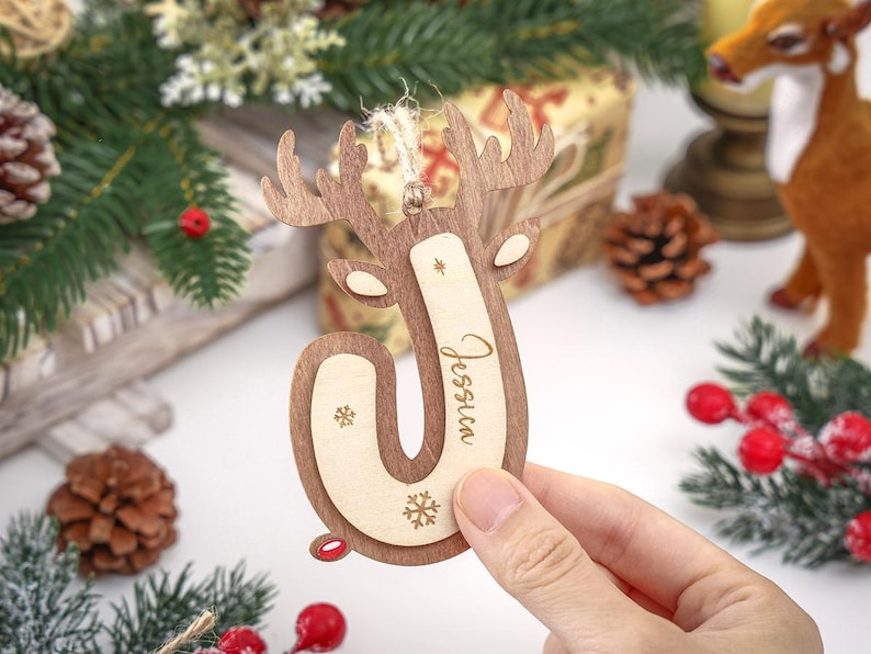 May include: A wooden Christmas ornament shaped like the letter J with reindeer antlers at the top. The ornament features a light wood center with the name Jessica engraved in script and two small snowflake icons, framed by a darker wood border.