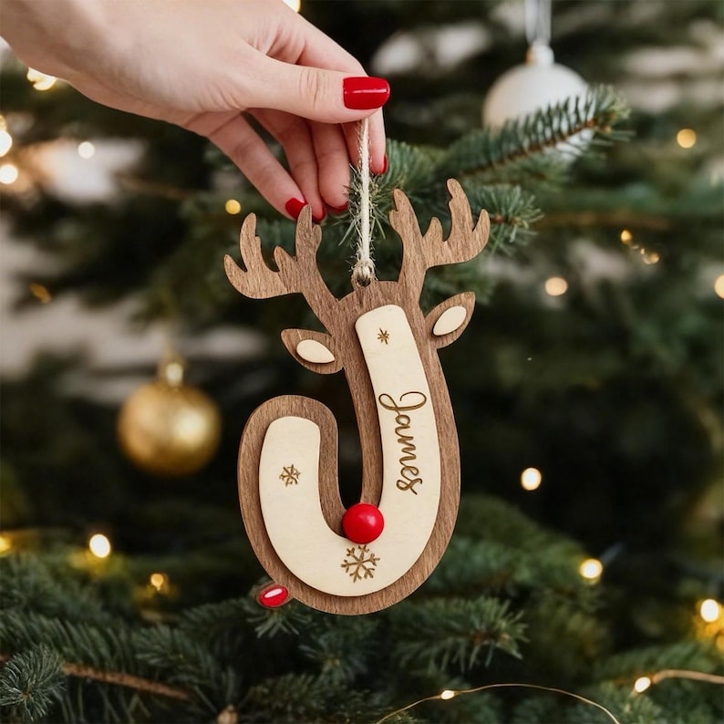 May include: A wooden reindeer Christmas ornament featuring a layered design with a light-colored wood insert engraved with the name James, a small red nose, and snowflake accents, suspended by a twine loop for hanging on a tree.