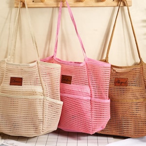 Puede incluir: Tres bolsos de malla en beige, rosa y marrón cuelgan de una pared de color claro. Cada bolso tiene un bolsillo rectangular en la parte delantera y una etiqueta de cuero con un nombre. Los bolsos tienen correas largas.