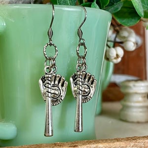 May include: A pair of silver-toned dangle earrings featuring a baseball glove and a baseball bat charm. The charms are suspended from a textured silver ring and a thin metal hook. The design is metallic with a polished finish and intricate details.
