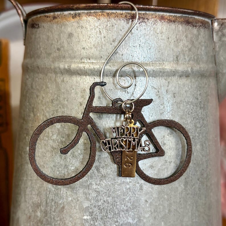 May include: A dark brown metal bicycle ornament featuring a silver-toned charm with the text Merry Christmas and a small rectangular tag with the number 26. The ornament includes a silver wire hook for hanging, designed with a spiral top loop.