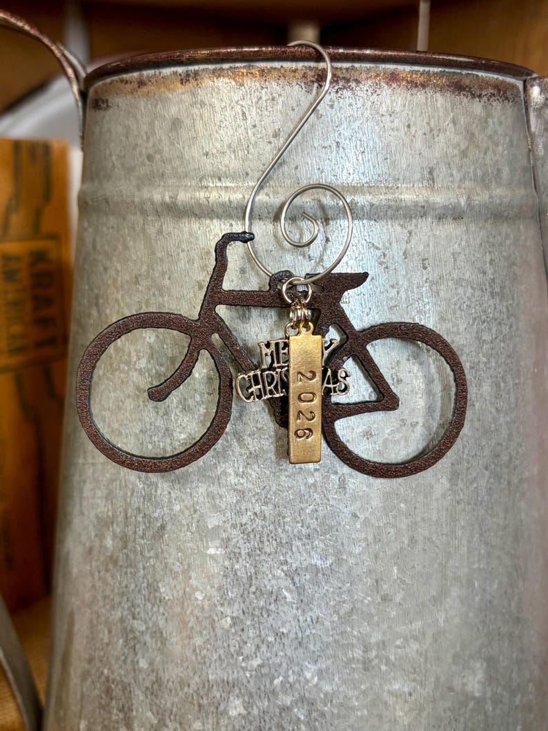 May include: A dark brown bicycle-shaped ornament hanging from a silver spiral hook. Attached to the frame is a silver charm with the text Merry Christmas and a vertical gold-toned rectangular charm engraved with the year 2026.