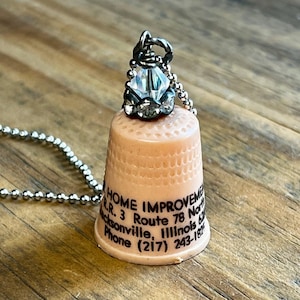 May include: A peach-colored plastic thimble pendant on a silver-toned ball chain. The thimble features black printed text reading HOME IMPROVEMENT, R.R. 3 Route 78 North, Jacksonville, Illinois 62650, Phone (217) 243-1824. A clear crystal bead sits on top.
