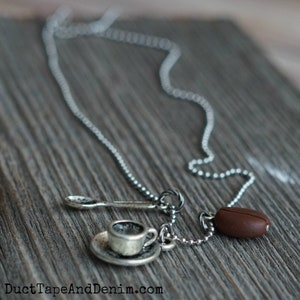 K&ouml;nnte beinhalten: Eine silberne Kettenkette mit einem Anh&auml;nger aus einer Kaffeetasse und Untertasse, einem L&ouml;ffel und einer Kaffeebohne.