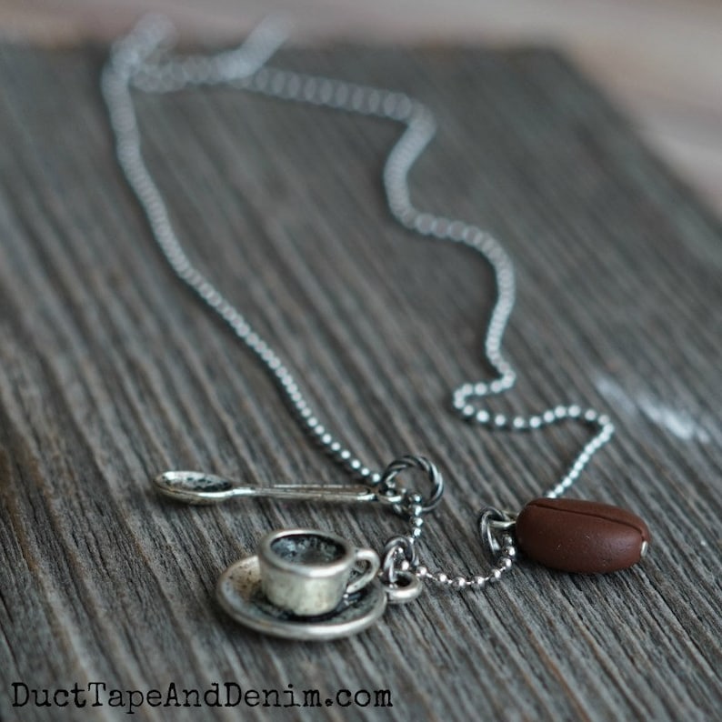 K&ouml;nnte beinhalten: Eine silberne Kettenkette mit einem Anh&auml;nger aus einer Kaffeetasse und Untertasse, einem L&ouml;ffel und einer Kaffeebohne.