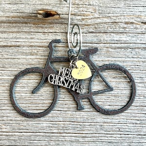 May include: A dark, textured metal bicycle ornament featuring a silver-toned wire hanger. Attached to the frame are two charms: a silver-colored script charm that reads Merry Christmas and a small, gold-toned heart charm engraved with 2024.