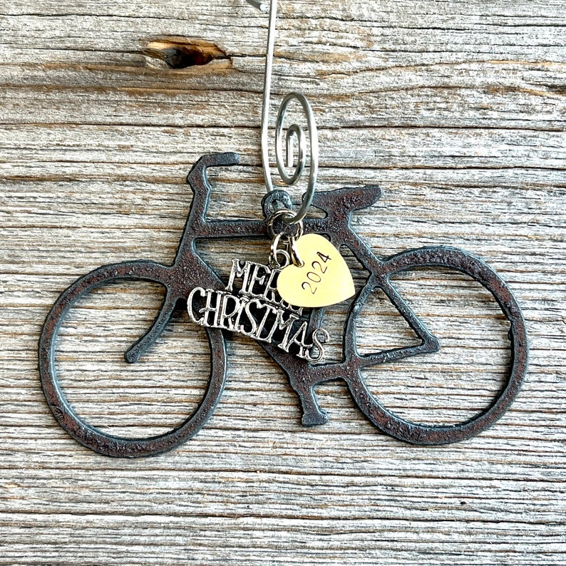 May include: A dark, textured metal bicycle ornament featuring a silver-toned wire hanger. Attached to the frame are two charms: a silver-colored script charm that reads Merry Christmas and a small, gold-toned heart charm engraved with 2024.