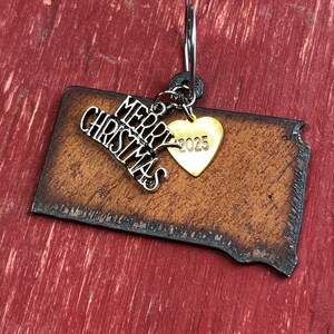 May include: A rectangular brown leather ornament with a dark distressed edge, featuring a silver-toned metal charm with the text Merry Christmas and a small gold-toned heart-shaped charm engraved with the year 2025, all attached to a metal ring.