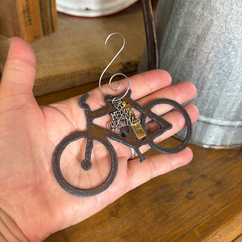 May include: A dark brown, rusted metal bicycle ornament featuring a silver wire hook at the top. A small, rectangular gold-toned metal charm hangs from the frame, engraved with the text Merry Christmas and the number 26 in a vertical orientation.