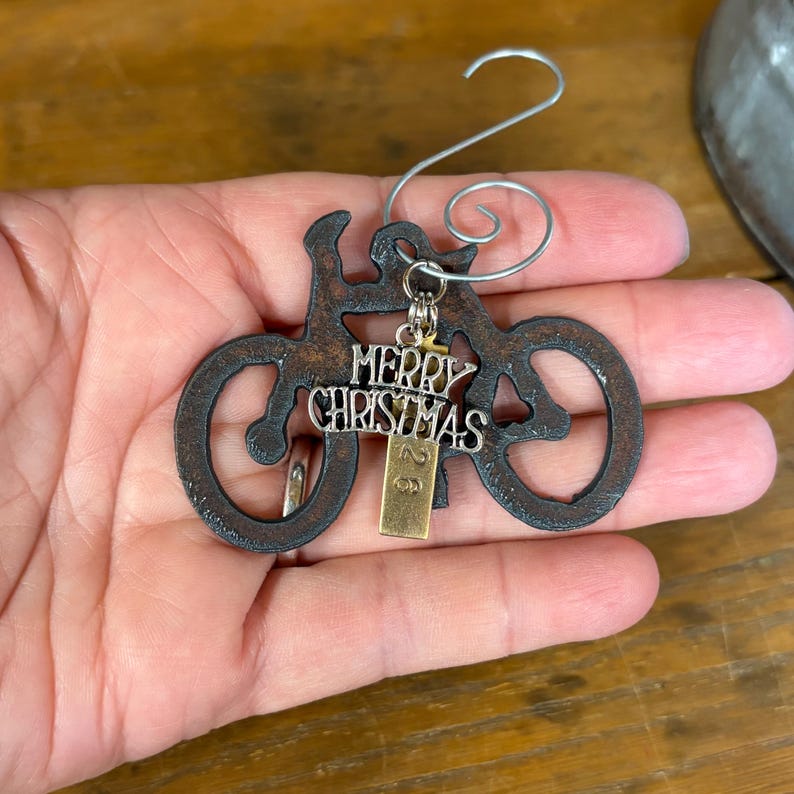 May include: A dark, rusted metal bicycle ornament featuring a silver-toned charm with the text Merry Christmas and a small rectangular brass-colored tag with the number 26. A silver wire hook is attached to the top of the bicycle frame.