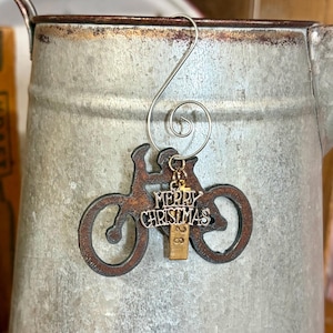 May include: A dark brown, rust-textured metal bicycle ornament featuring a silver-toned charm with the text Merry Christmas and a small rectangular gold-toned tag with the number 26, all suspended from a silver-toned metal spiral wire hook.