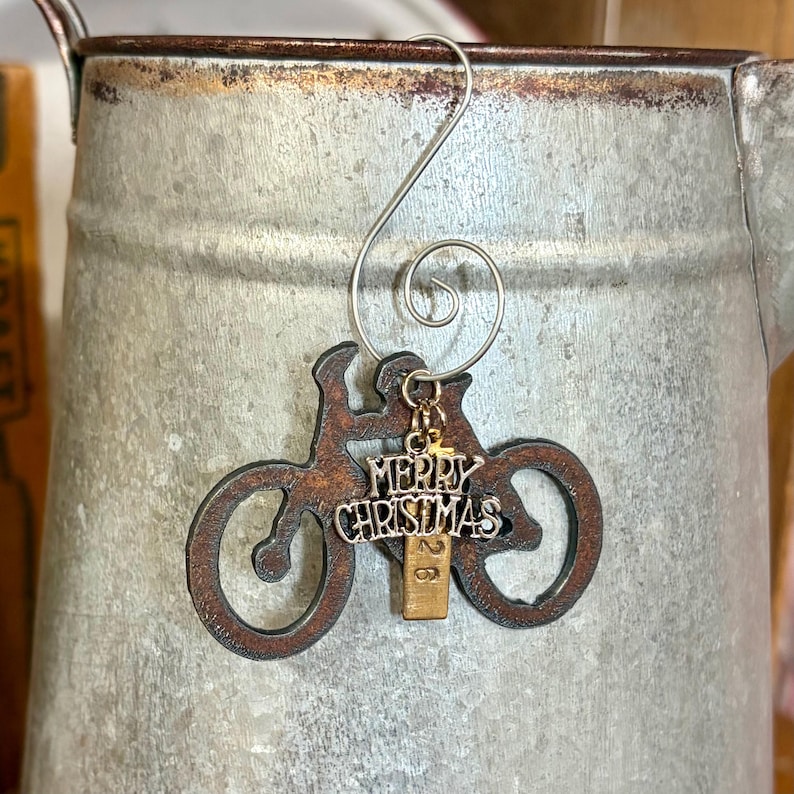 May include: A dark brown, rust-textured metal bicycle ornament featuring a silver-toned charm with the text Merry Christmas and a small rectangular gold-toned tag with the number 26, all suspended from a silver-toned metal spiral wire hook.