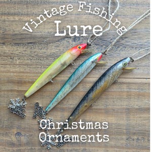May include: Three fishing lure Christmas ornaments with twine hangers. The lures are yellow with a red head, teal with a silver body, and black with a gold body. Each features a silver snowflake charm with the text Merry Christmas attached.