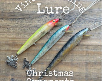 May include: Three fishing lure Christmas ornaments with twine hangers. The lures are yellow with a red head, teal with a silver body, and black with a gold body. Each features a silver snowflake charm with the text Merry Christmas attached.