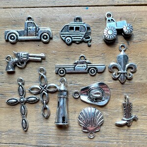 May include: A collection of eleven silver-toned metal charms featuring various shapes including a pickup truck, a camper trailer, a tractor, a revolver, a sedan, a fleur-de-lis, two different cross designs, a lighthouse, a cowboy hat, a seashell, and a boot.