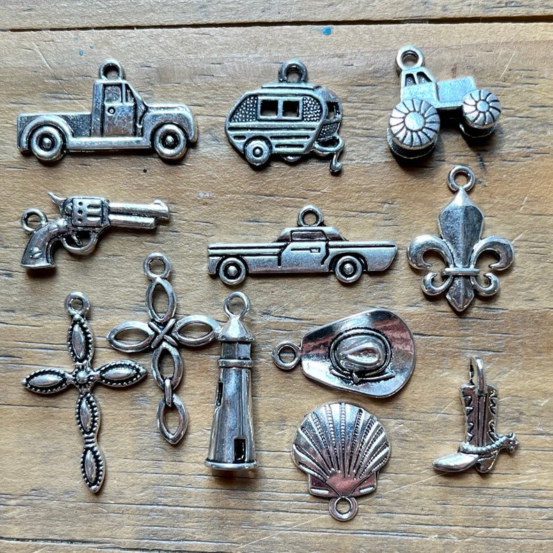 May include: A collection of eleven silver-toned metal charms featuring various shapes including a pickup truck, a camper trailer, a tractor, a revolver, a sedan, a fleur-de-lis, two different cross designs, a lighthouse, a cowboy hat, a seashell, and a boot.