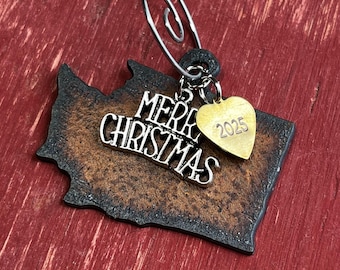 May include: A state-shaped ornament with a dark, textured finish featuring a silver-toned metal charm that reads Merry Christmas and a small gold-toned heart charm engraved with the year 2025, all suspended from a silver wire hanging loop.