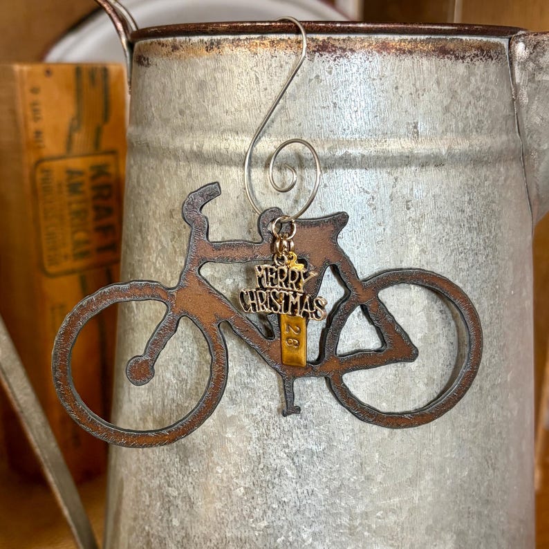 May include: A rust-colored metal bicycle ornament featuring a silver-toned wire hanger and a small gold-toned charm dangling from the frame. The charm displays the text Merry Christmas and the number 28 in a vertical orientation on a rectangular tag.