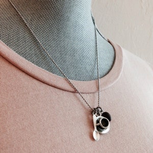 K&ouml;nnte beinhalten: Eine silberne Kettenkette mit einem Anh&auml;nger in Form einer kleinen Kaffeetasse, Untertasse und L&ouml;ffel. Der Anh&auml;nger h&auml;ngt an einer silbernen Kette.