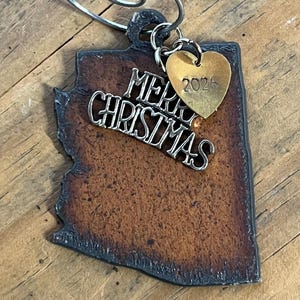 May include: A dark brown, textured, Arizona-shaped ornament with a dark metal border. It features a silver-toned metal script cutout reading Merry Christmas and a small, gold-toned heart-shaped charm stamped with the year 2024 attached by a ring.
