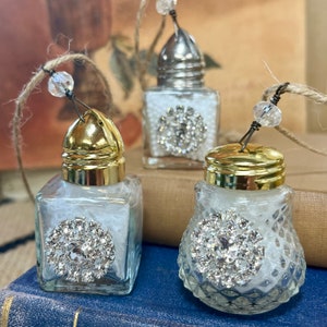 May include: Three glass salt and pepper shakers with gold-tone metal lids, each featuring a circular rhinestone embellishment on the front and a clear crystal bead attached to a jute twine loop for hanging. The glass containers are filled with white material.