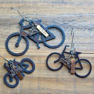 May include: Three bicycle-shaped ornaments with a dark, rusted metal finish. Each ornament features a silver-toned charm with the text Merry Christmas and a rectangular metal tag stamped with the year 2026, suspended by a curved metal wire hook.