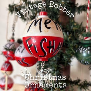 May include: A spherical fishing bobber Christmas ornament with a white top and red bottom. The text "ME" is painted in black on the white section, and "FISHY" is painted in black on the red section. A silver-toned metal fish charm hangs below.