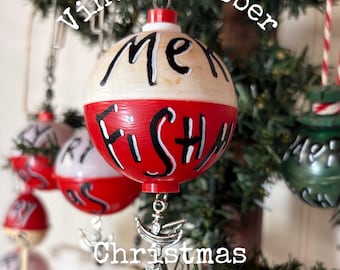 May include: A spherical fishing bobber ornament painted red and white with the hand-painted black text "ME" on the top and "FISHY" on the bottom. A silver-toned metal fish hook charm hangs from the bottom, and a metal loop is at the top.