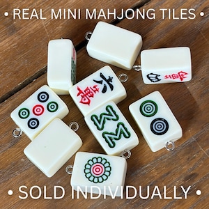 May include: A collection of off-white mini Mahjong tiles, each featuring a small metal jump ring attached for use as a charm. The tiles display various traditional symbols, characters, and floral patterns in red, green, and black ink. Text reads: REAL MINI MAHJONG TILES SOLD INDIVIDUALLY.